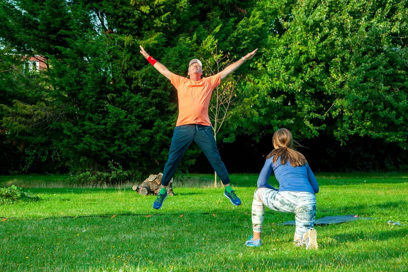 Man exercising doing a star jump with Personal Trainer outside in the park